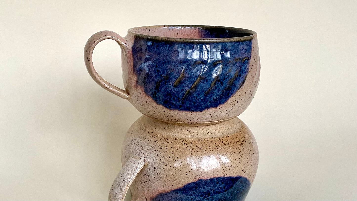 A generously sized soup mug with a wide, low profile, but plenty of depth. The mouth comes in slightly to counteract sloshing or spilling. A comfortable handle attaches to one side and features gentle contouring on the underside ensuring an easy grip. Glaze options include a deep indigo atop a shell pink, or a rich blue-green verdigris atop a warm sandy golden brown.